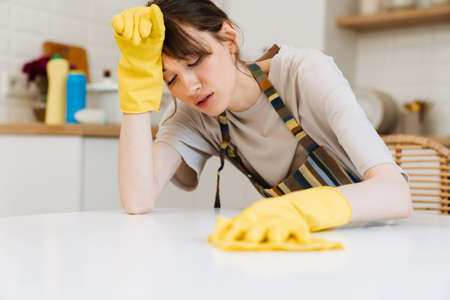 White young woman in gloves wiping her forehead while cleaning table at homeの写真素材