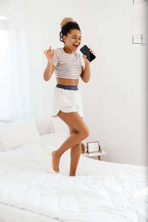 Young black woman singing while dancing with cellphone on bed at home