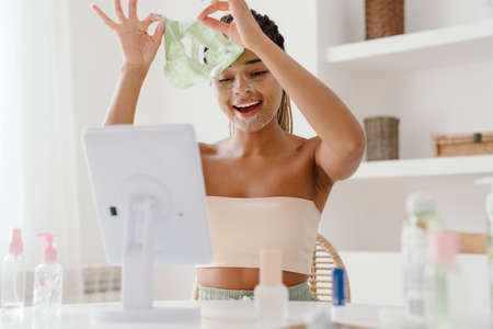 Young black woman with cosmetic mask looking in mirror at homeの写真素材