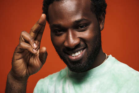 Young black man with beard gesturing and smiling at camera isolated over brown wall