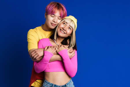 Asian couple with multicolored hair hugging while smiling together isolated over blue wallの写真素材