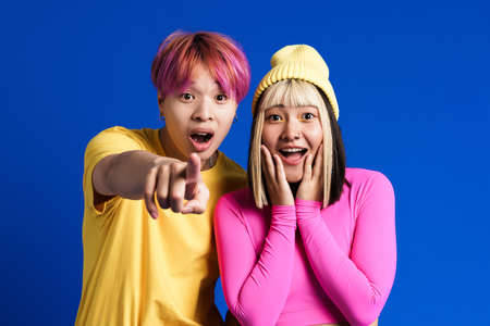 Asian teenagers pointing finger at camera while expressing surprise isolated over blue wall