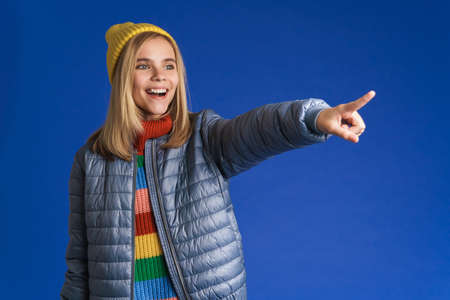 White girl wearing jacket smiling and pointing finger aside isolated over blue background