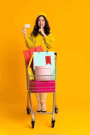 White woman posing with credit card and shopping cart isolated over yellow background