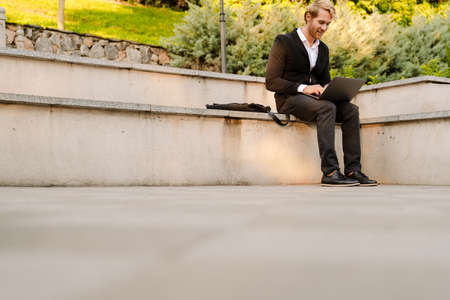 Blonde man working with laptop while sitting on stone bench outdoorsの写真素材