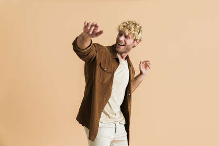 White blonde man wearing shirt smiling and dancing on camera isolated over beige background