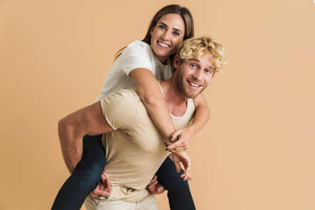 White couple wearing t-shirts smiling while piggybacking isolated over beige background