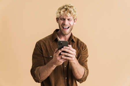 White blonde man wearing shirt laughing and using cellphone isolated over beige backgroundの写真素材