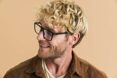 White blonde man in eyeglasses smiling and looking aside isolated over beige background