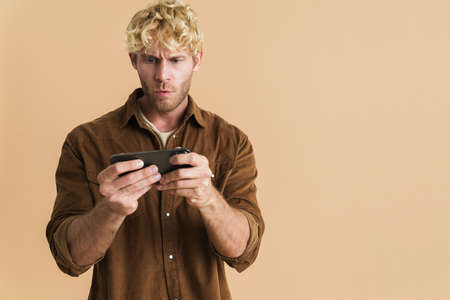 White blonde man wearing shirt playing online game on cellphone isolated over beige backgroundの写真素材