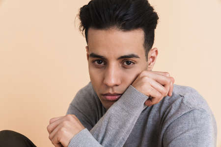Young middle eastern man looking at camera while sitting on floor isolated over beige background