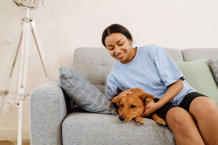 Young hispanic woman smiling and petting her dog while resting on couch at home