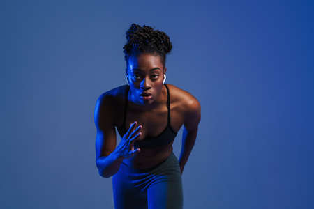 Black sportswoman in earphones doing exercise while working out isolated over blue backgroundの写真素材