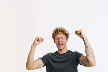 Young blonde man screaming while making winner gesture isolated over white wall