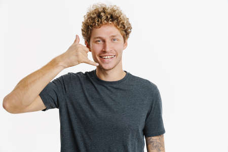 Young blonde man with curly hair smiling and making call gesture isolated over white wall