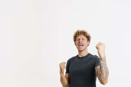 Young blonde man screaming while making winner gesture isolated over white wall