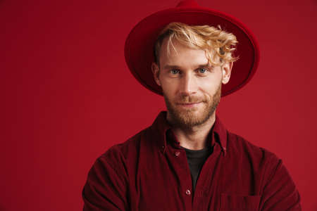 White bearded man wearing hat smiling and looking at camera isolated over red wall