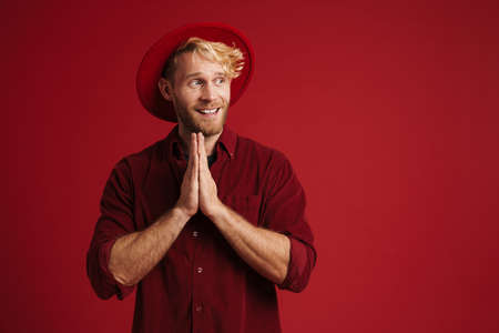White bearded man wearing hat smiling while making prayer gesture isolated over red wallの写真素材