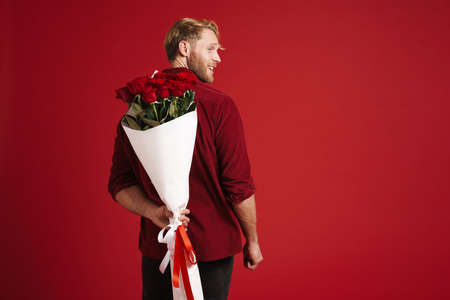 Blonde man smiling while posing with bouquet of roses isolated over red backgroundの写真素材