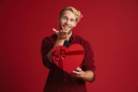 Blonde man holding copyspace while showing heart gift box isolated over red wallの写真素材