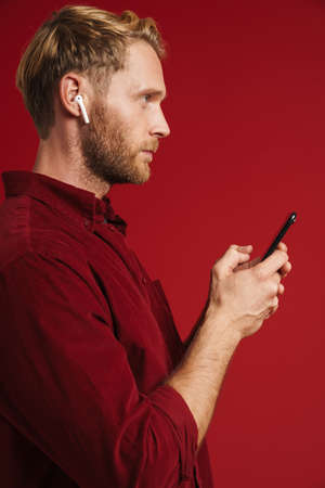 Blonde man listening music with earphones and using cellphone isolated over red wall