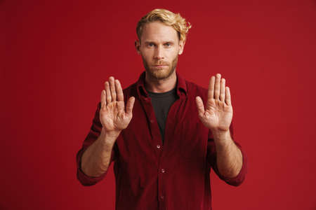 White bearded man wearing shirt showing stop gesture at camera isolated over red wall