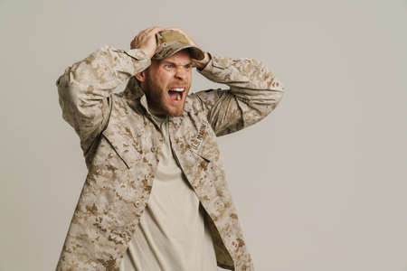 Furious military man wearing uniform holding head and screaming isolated over white wall
