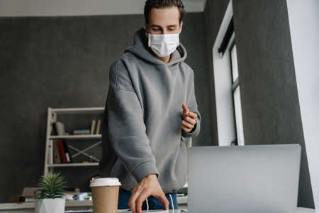 Young white man in protective mask working with laptop at officeの写真素材