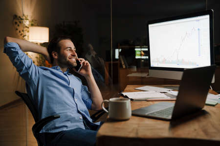 Young bristle man talking on mobile phone while working with laptop in officeの写真素材