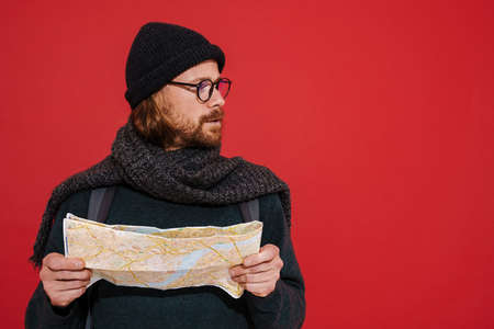 Ginger puzzlement man wearing hat and scarf examining map isolated over red backgroundの写真素材
