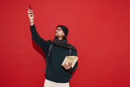 Ginger puzzlement man examining map and using mobile phone isolated over red background