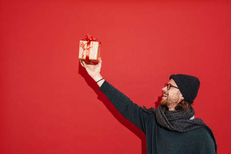 White man in warm clothes and eyeglasses showing gift box isolated over red background