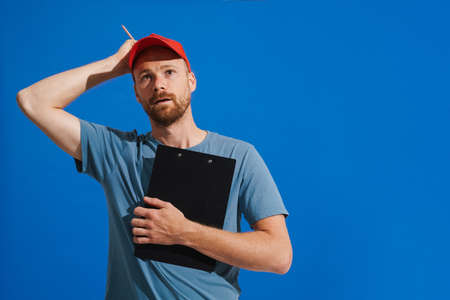 White puzzled man wearing cap looking upward while posing with clipboard isolated over blue...