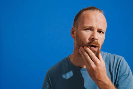Ginger puzzled man holding his chin while looking at camera isolated over blue background