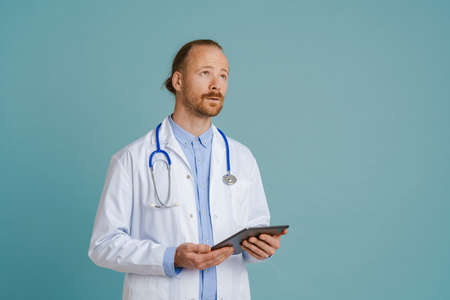 White male doctor wearing lab coat using tablet computer isolated over blue background