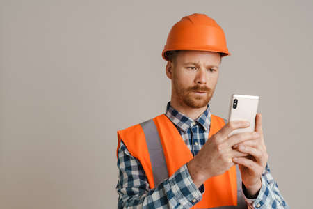 White man worker wearing helmet and vest using mobile phone isolated over grey background