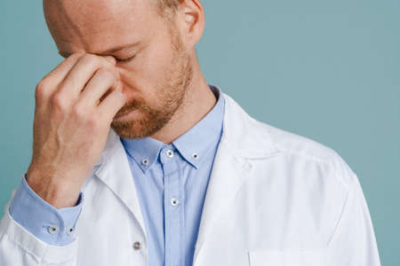 Tired male doctor with headache posing on camera isolated over blue background
