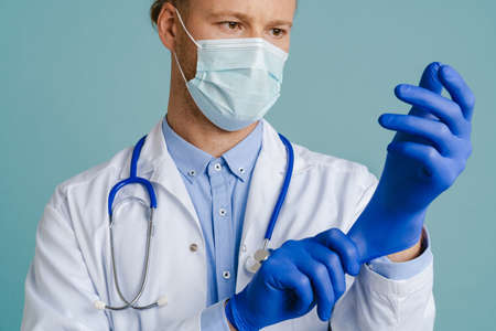 White male doctor in face mask wearing medical gloves isolated over blue backgroundの写真素材