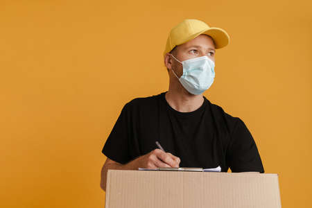 White delivery man in face mask writing down notes on clipboard isolated over yellow backgroundの写真素材