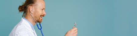 White male doctor wearing lab coat smiling and using stethoscope isolated over blue backgroundの写真素材