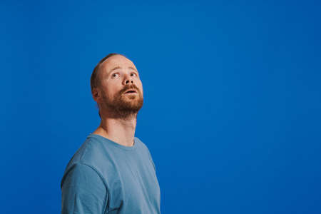 Ginger puzzled man with beard posing and looking upward isolated over blue background