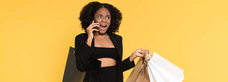 Black woman talking on cellphone while posing with shopping bags isolated over yellow background