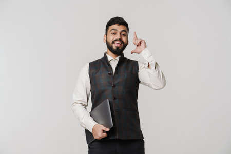 Bearded south asian man pointing finger upward while posing with laptop isolated over white wall