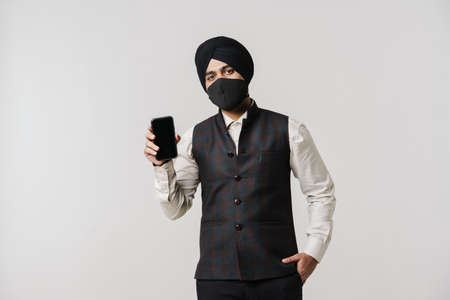 South asian man wearing turban and face mask showing cellphone isolated over white wall