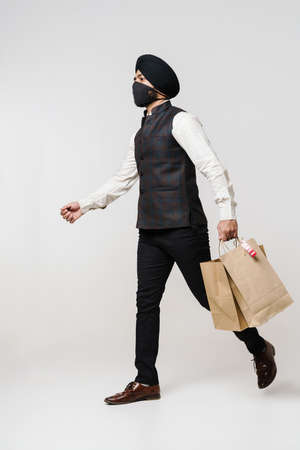 Bearded south asian man in face mask walking with shopping bags isolated over white wallの写真素材