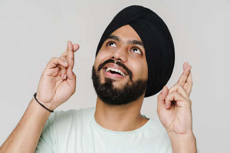 Bearded south asian man wearing turban holding fingers crossed isolated over white wallの写真素材