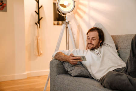 Ginger bristle man using mobile phone while resting on couch at homeの写真素材