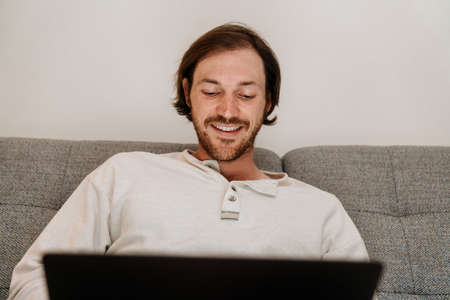 European unshaven man using laptop while resting on sofa at homeの写真素材