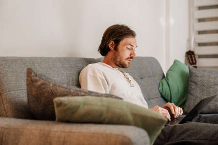 European unshaven man using laptop while resting on sofa at home