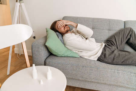 Ginger man feeling sick and covering his face while lying on couch at homeの写真素材